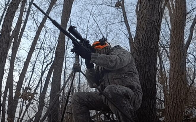A man in camo sits and aims his airgun that is on a tripod